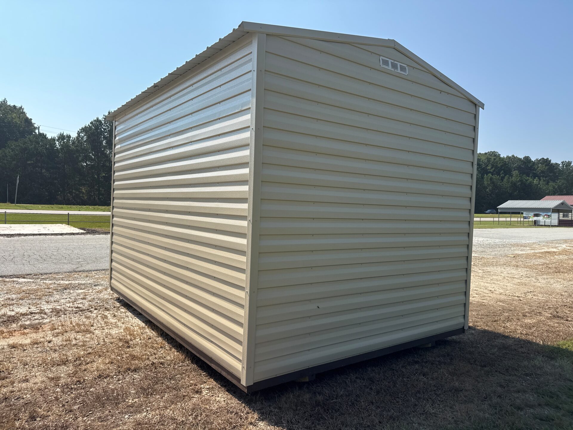 10x14 beige metal Estate Garage Shed with sloped roof on dry grass near road, trees and building in back, clear blue sky above.