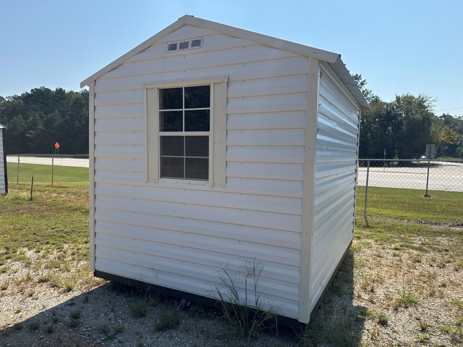 8x10 white metal Ridgeline Shed with window and vent on gravel near chain-link fence, grass, trees, and sun. Available now.