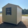 The 8x8 Ridgeline Shed (#082541439), beige with dark windows, sits on dry grass by a road, trees, and fence under clear skies.