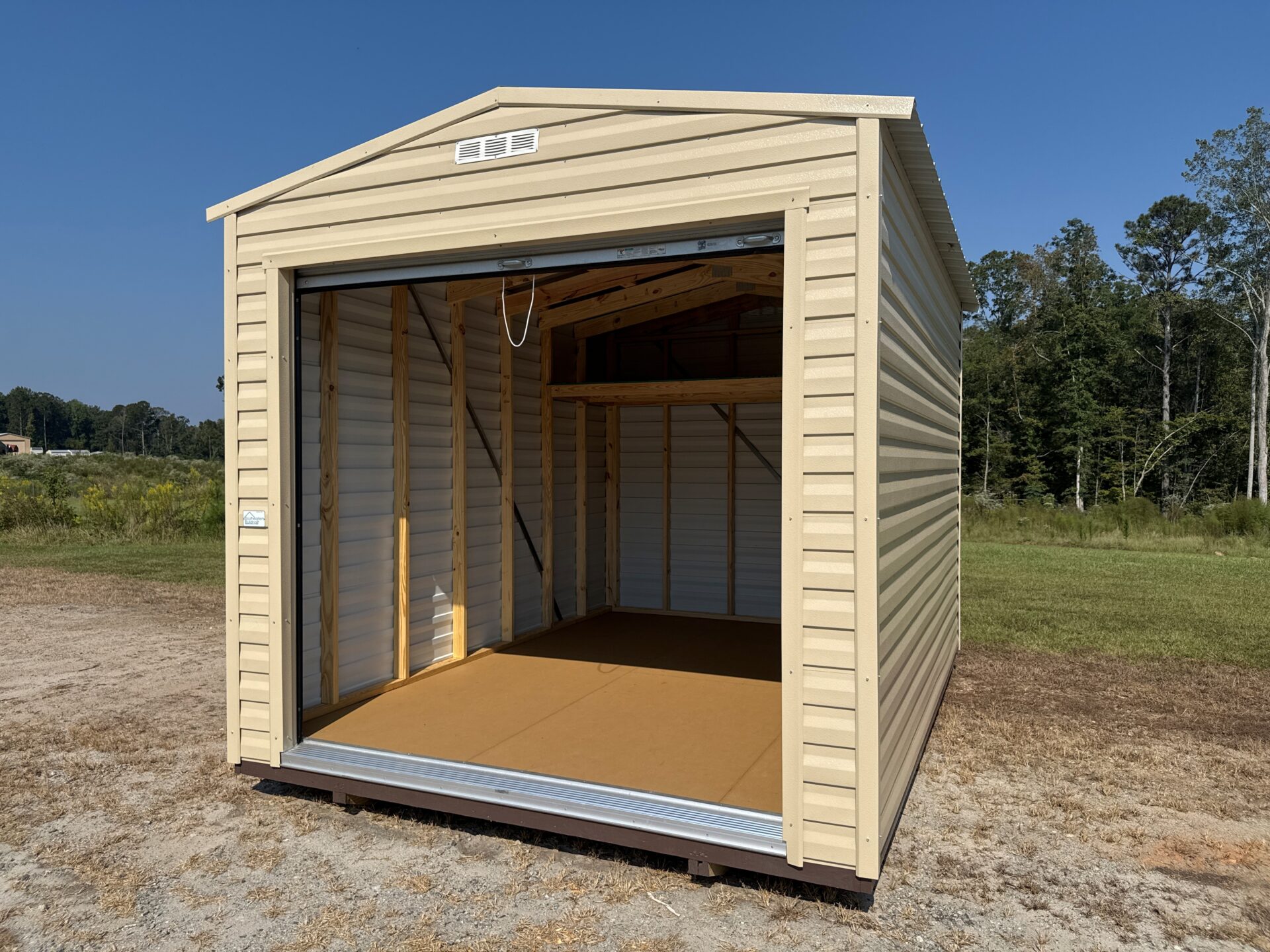 A tan 10x14 garage shed with an open roll-up door sits on dry grass, empty inside. Trees and blue sky are in the background.
