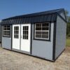 The 12x16 Frontier Shed with gray exterior, white double doors, two windows, black metal roof sits on gravel among grass and trees.