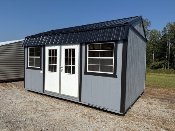 The 12x16 Frontier Shed with gray exterior, white double doors, two windows, black metal roof sits on gravel among grass and trees.