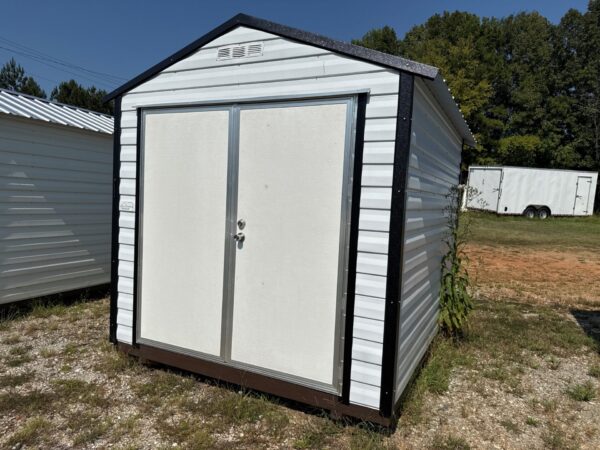 An 8x10 Scratch and Dent Ridgeline Shed with double doors and dark roof sits on grass; trailer behind, sunny sky. Available now.