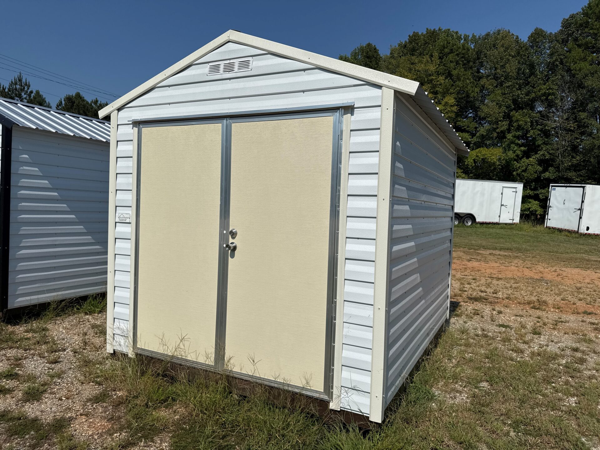 The 8x10 Scratch and Dent - Ridgeline Shed is a small white metal shed with double doors and peaked roof on grass, trees behind.