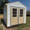 An 8x10 white Ridgeline Shed with double doors and two windows stands on grass, trees and buildings behind. Stock #102840945.