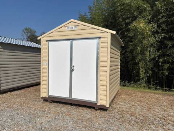 The 8x8 Ridgeline Shed (#082541439), a beige metal shed with white double doors and sloped roof, stands on dry grass and gravel.