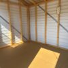 Interior of empty 8x12 Ridgeline Shed 022041155 with wooden framing, beige floor, white metal walls and roof, sunlight casting a shadow.