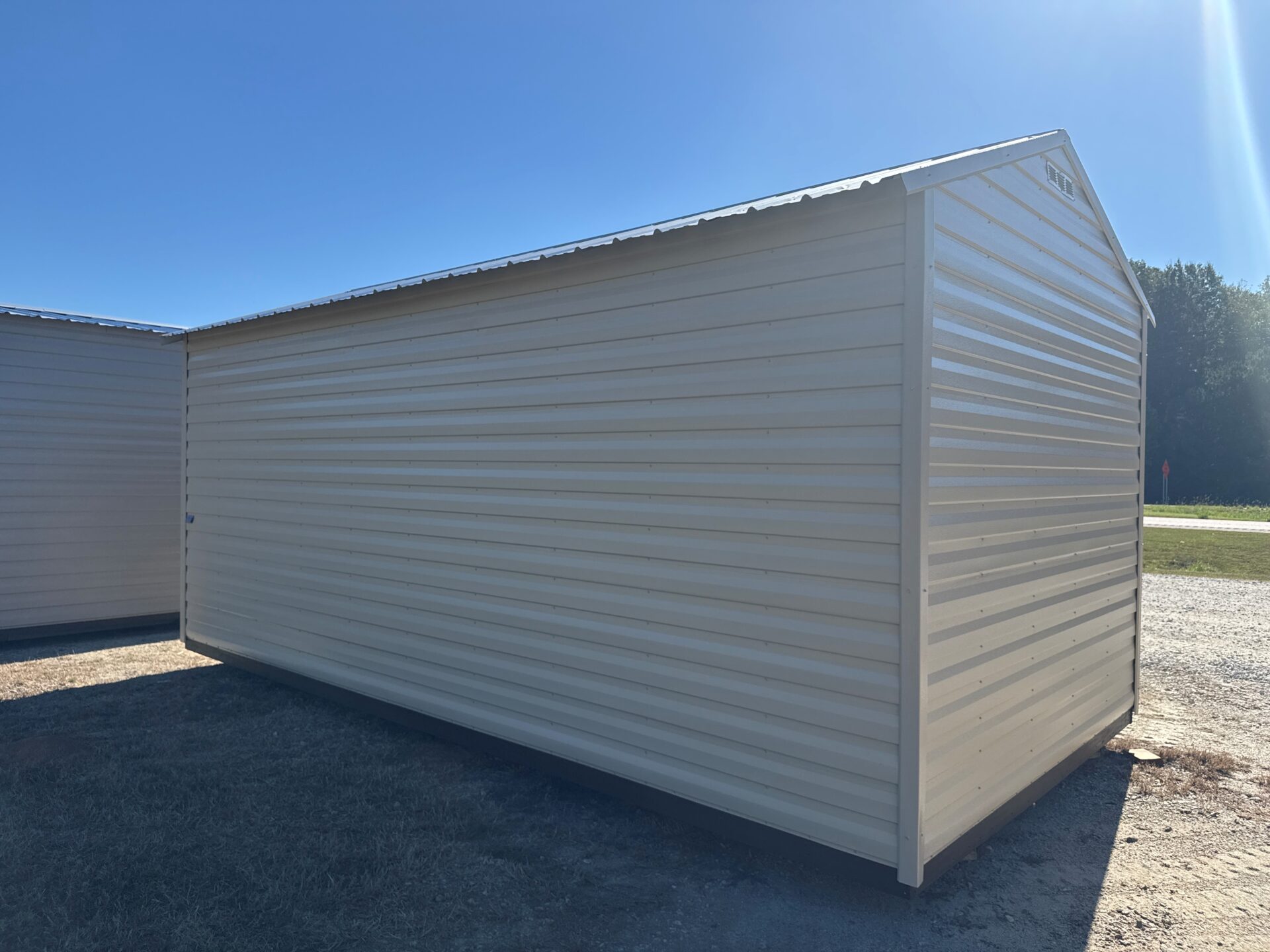 The 10x20 Ridgeline Garage Shed with beige ribbed metal siding and a sloped roof sits on gravel, sunlight, trees, and road behind.