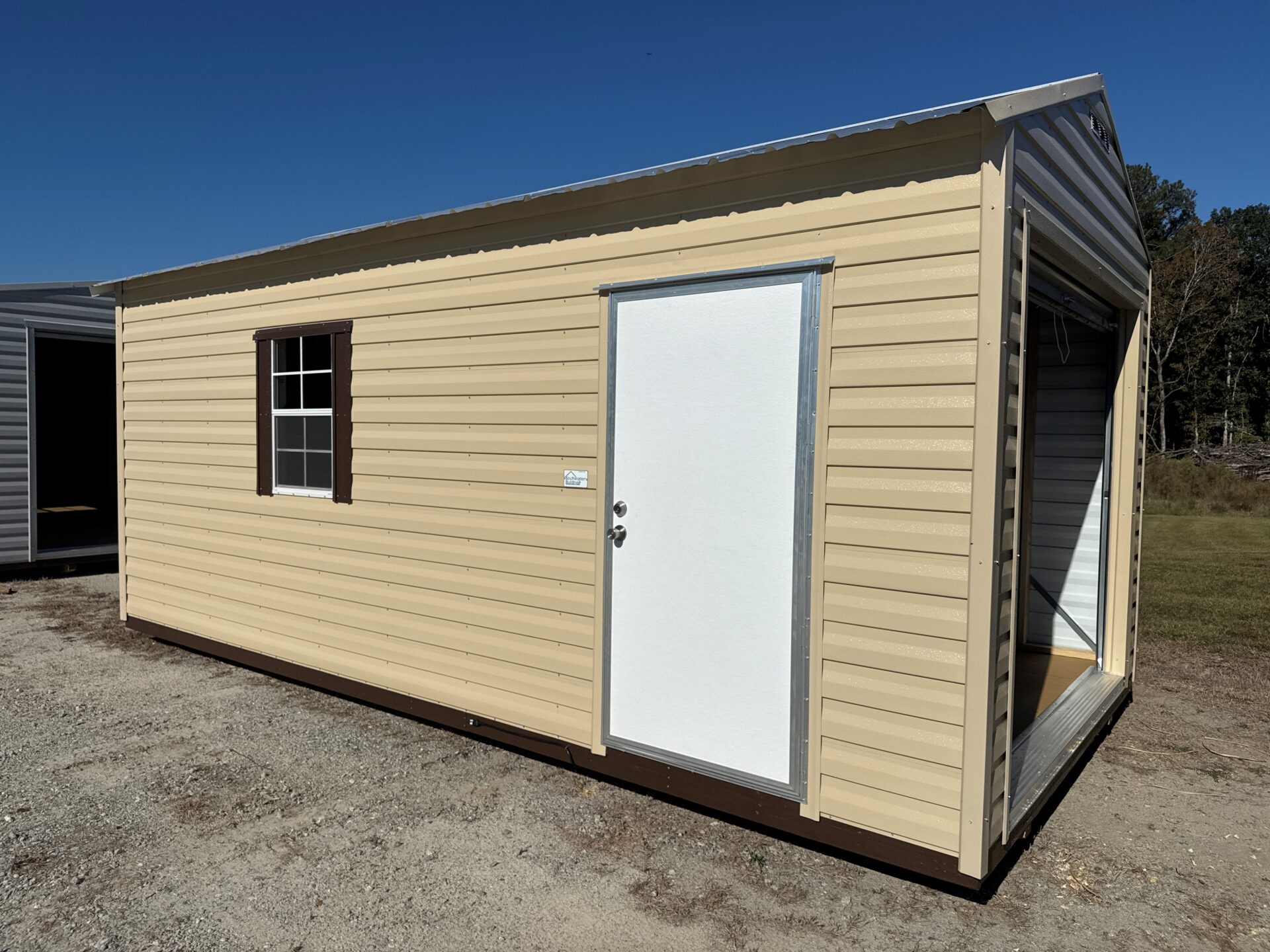 The 10x20 Ridgeline Garage Shed features beige metal siding, a white door, window with dark shutters, and open garage door.