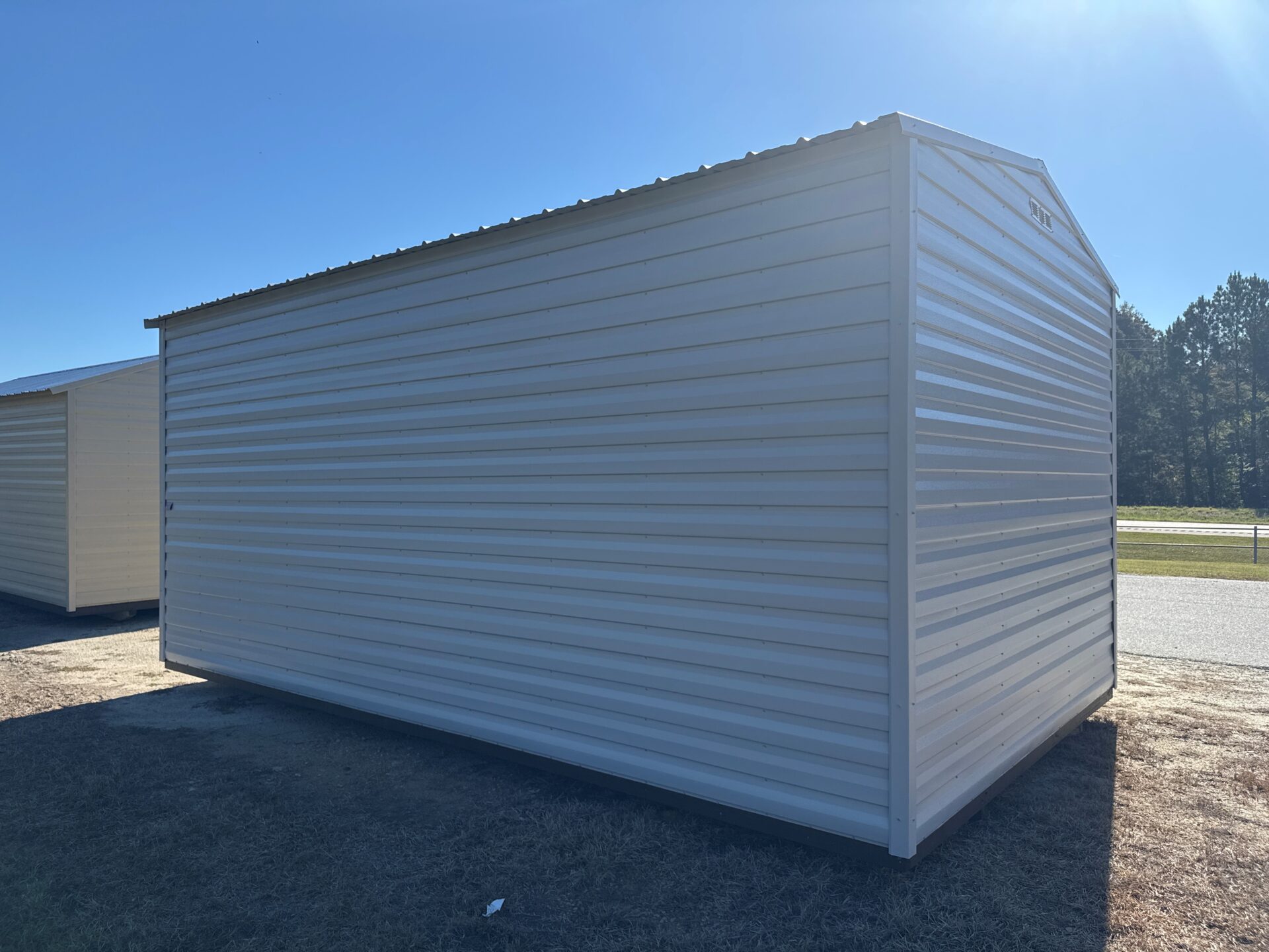 A 10x16 Estate Garage Shed Stock #082541440 with light gray metal and sloped roof sits on dry grass beside a road, clear blue sky.