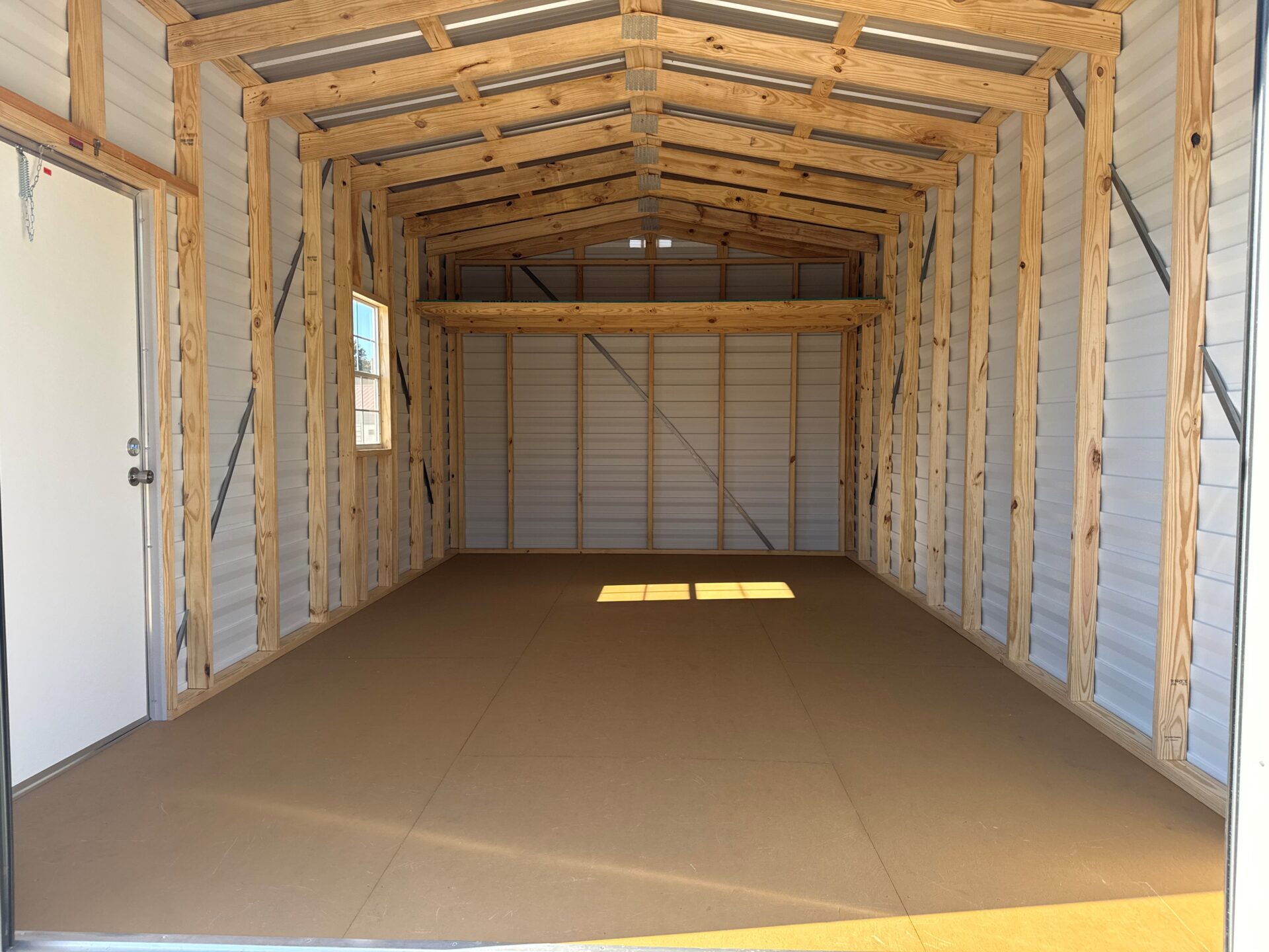 Interior of the 12x20 Estate Garage Shed with exposed beams, metal roof, left-side window, closed white door, sunlight on floor.