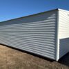 A 12x28 Ridgeline Garage Shed, white ridged metal exterior, on dry grass by a road under a clear blue sky. Available now.