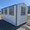 The 10x16 Frontier Shed, stock #0411M41250, with white siding, gray trim, and 3 windows on gravel near trees under blue sky.