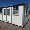 The 10x16 Frontier Shed is a small white shed with gray metal roof, glass double doors, and four windows on a gravel lot.