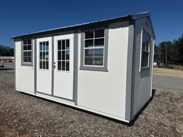 The 10x16 Frontier Shed is a small white shed with gray metal roof, glass double doors, and four windows on a gravel lot.