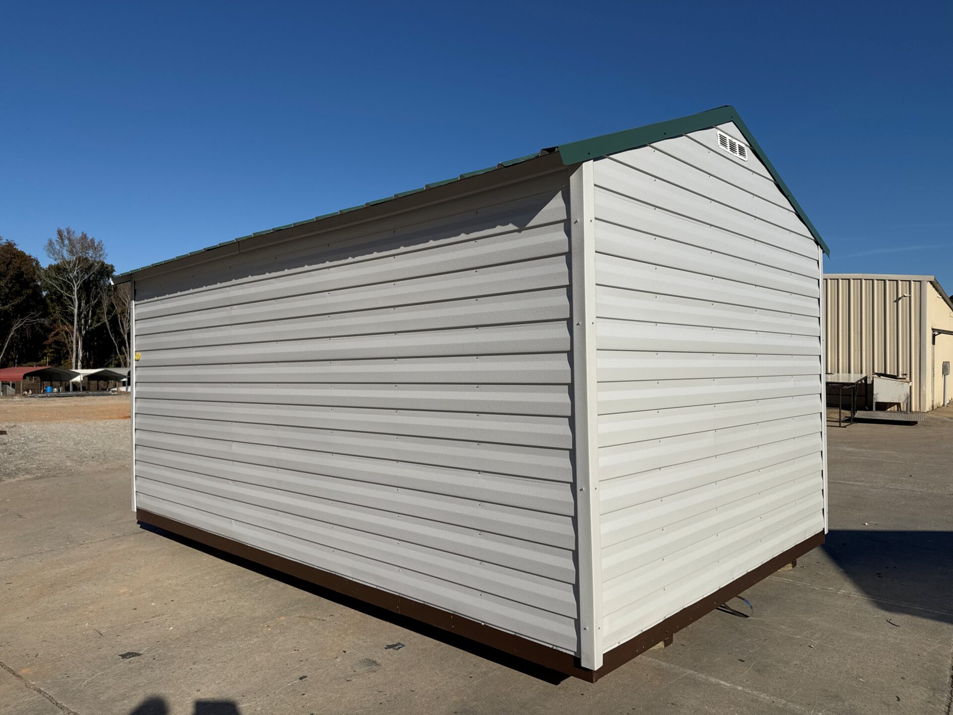 The 10x16 Ridgeline Shed with metal siding and a green roof sits on a concrete pad outdoors near other buildings and trees.