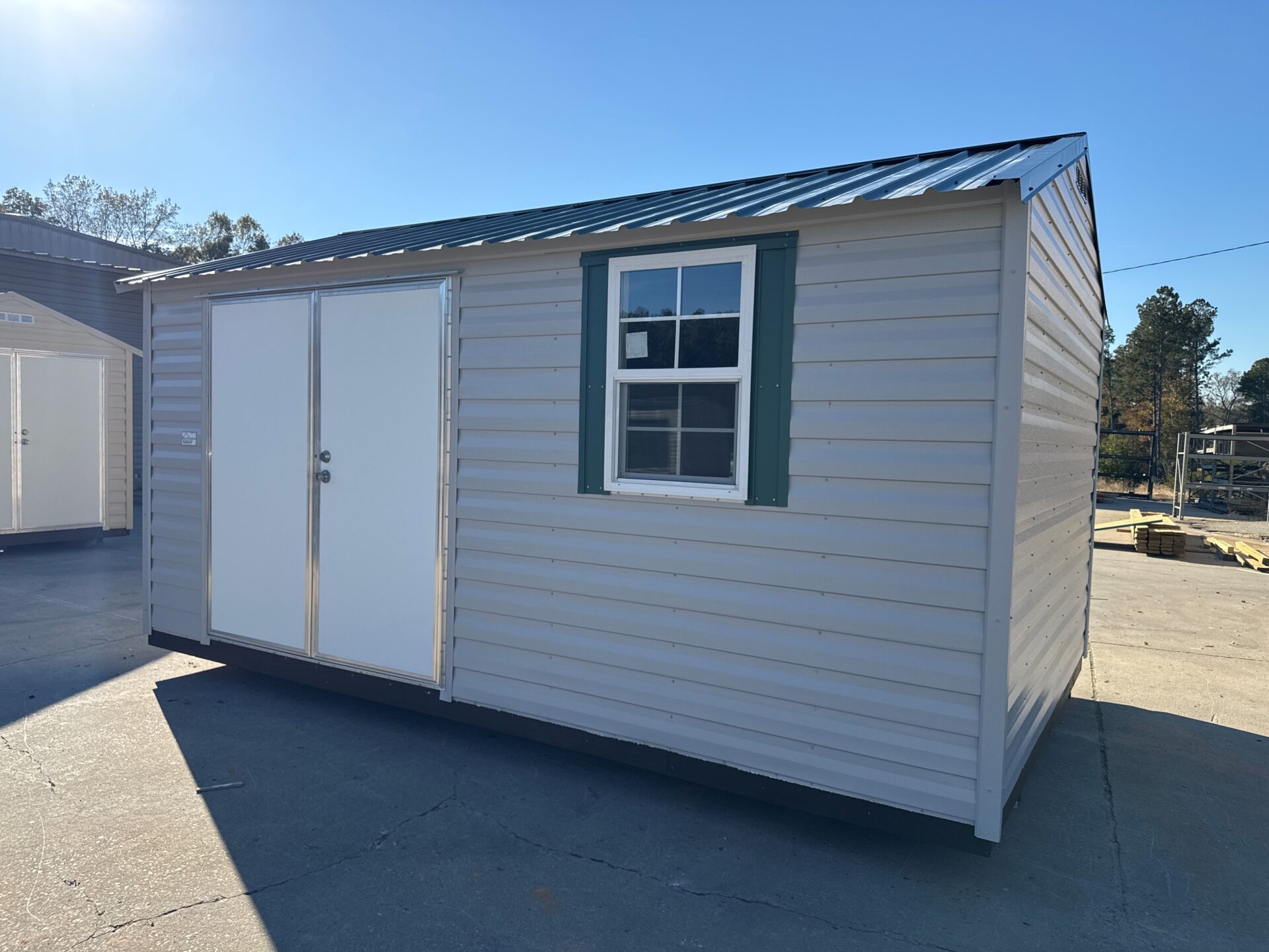 The 10x16 Ridgeline Shed, light gray with a green-trimmed window and white double doors, sits on a concrete lot under blue sky.