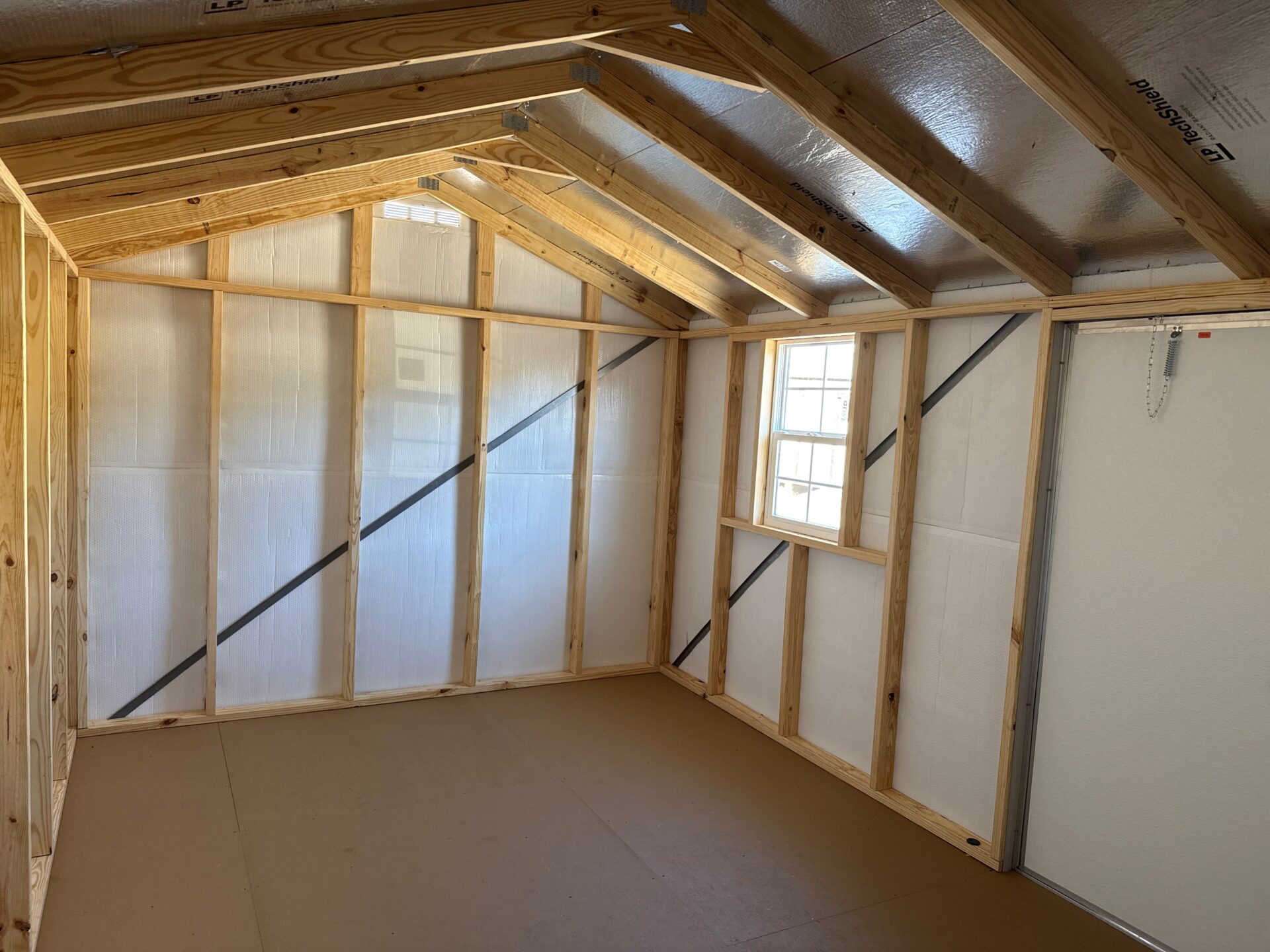 Interior of the 10x16 Ridgeline Shed shows exposed wooden framing, a sloped insulated roof, single window, bare floor, and door.