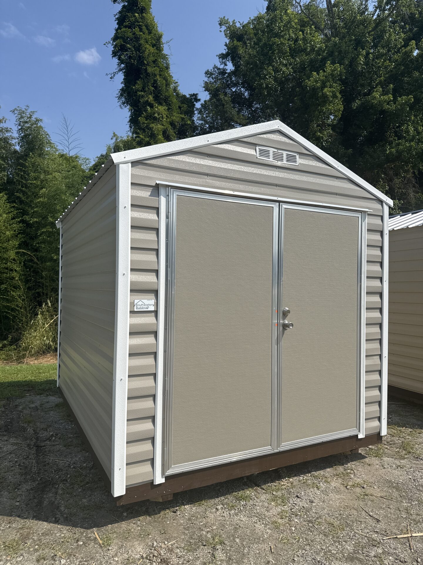 An 8x10 Ridgeline Shed with Hickory Moss metal exterior, double doors, and silver frame stands on dirt and grass amid trees under a blue sky.
