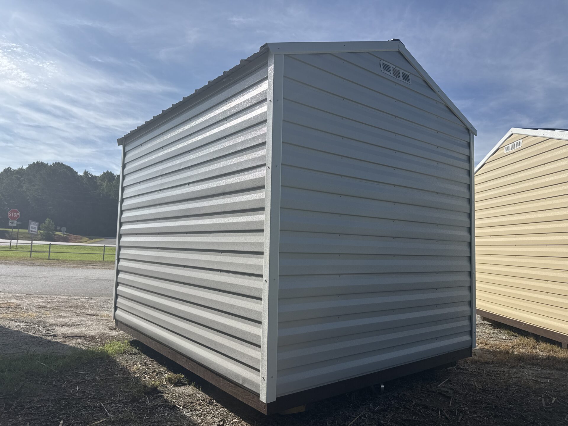 An 8x10 Ridgeline Shed (Stock #041341716), featuring a small, white, metal design with a gable roof, is situated on a patch of dirt and grass near a road, with trees and another yellow shed visible in the background beneath a blue sky.