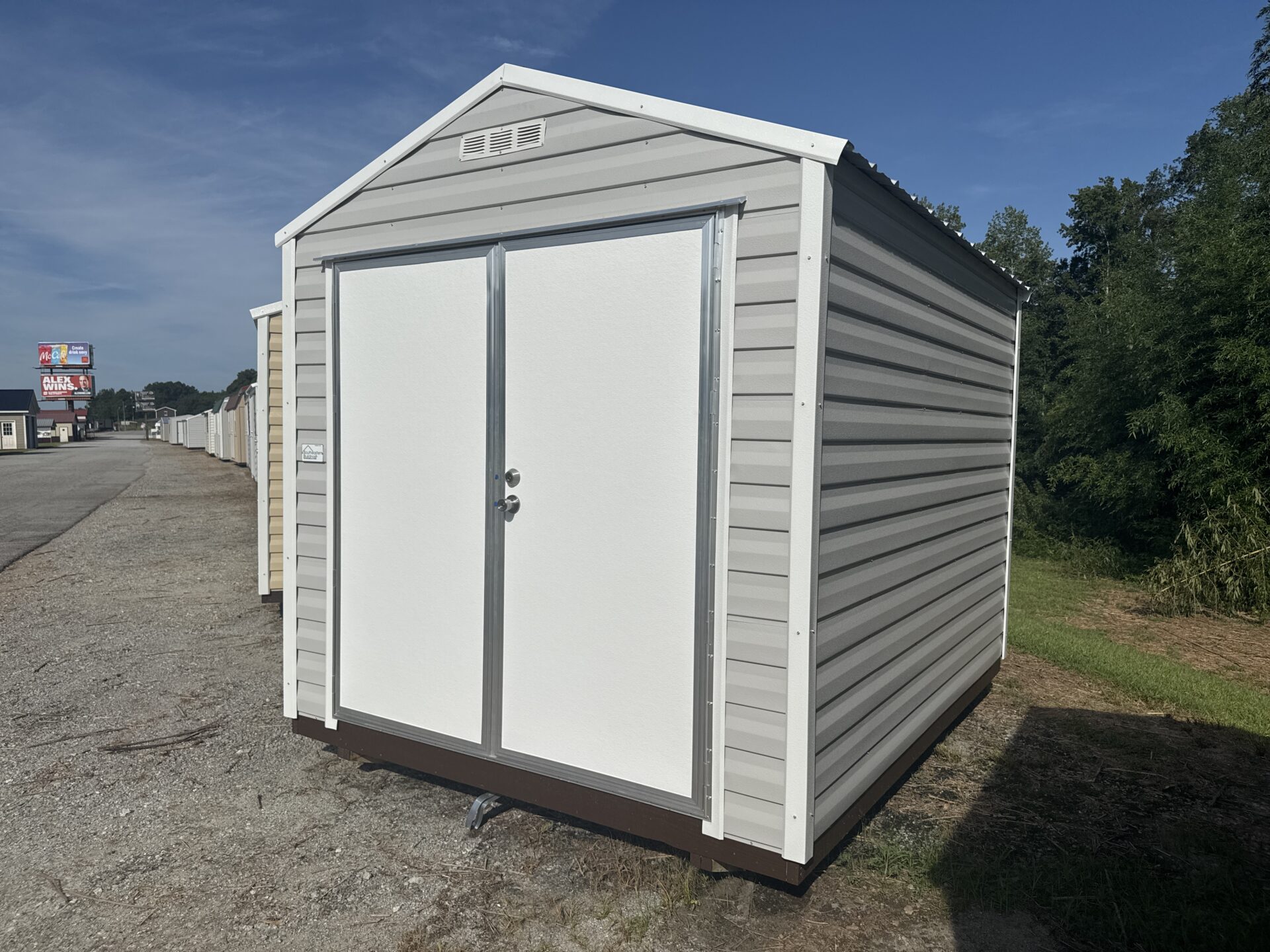 An 8x10 Ridgeline Shed (Stock #071641798), featuring a compact white metal design with double doors, is set on gravel near a grassy area, with trees and other sheds visible in the background beneath a clear blue sky.