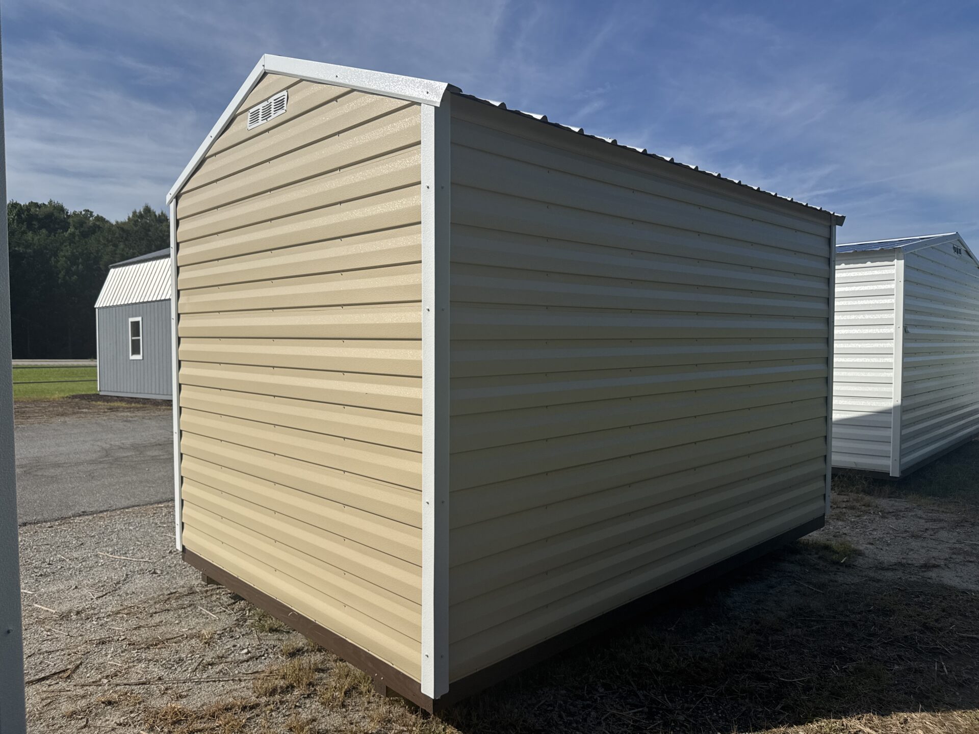 An 8x12 Ridgeline Shed (Stock #071341792), featuring a beige metal exterior with a white roof and trim, sits on gravel and grass. Similar sheds are visible in the background beneath a blue sky.