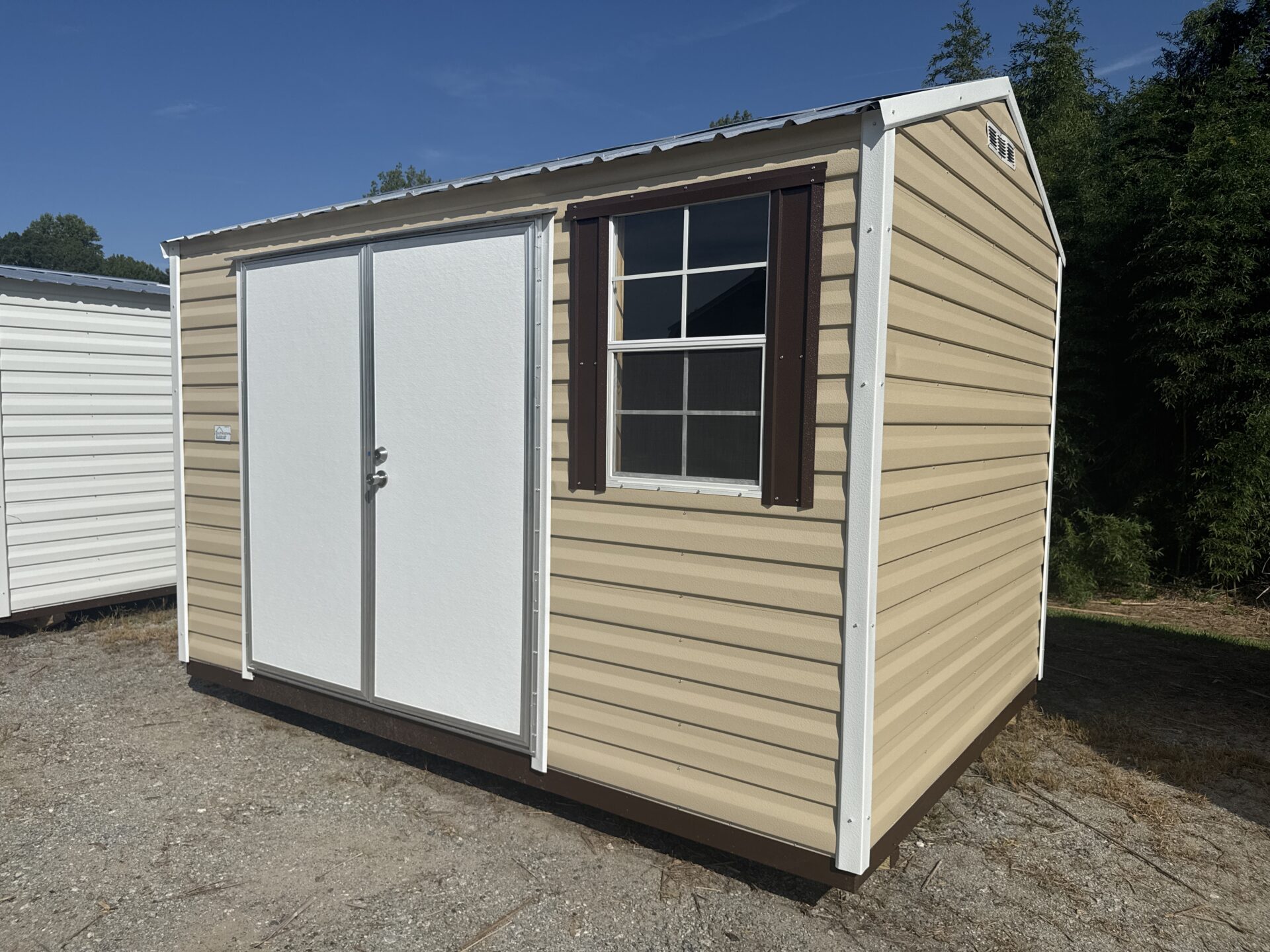 The 8x12 Ridgeline Shed - Available Now (Stock #071341792) features a small beige exterior with a metal roof, double white doors, and a window with dark brown shutters, and is situated on a gravel lot with trees and another white shed in the background.