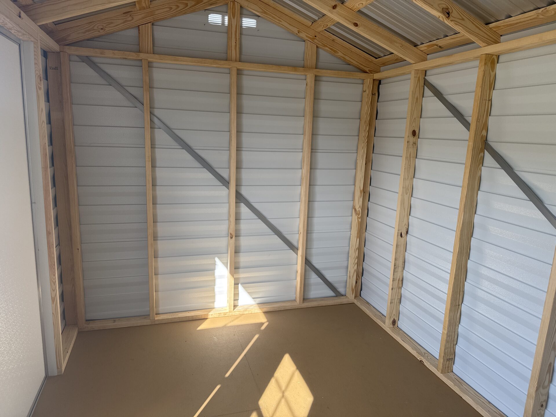 The interior of the 8x12 Ridgeline Shed (Stock #071341792) features unfinished wooden framing, metal walls and roof, and a simple brown floor. Sunlight streams through a window, casting shadows on the wall and floor.