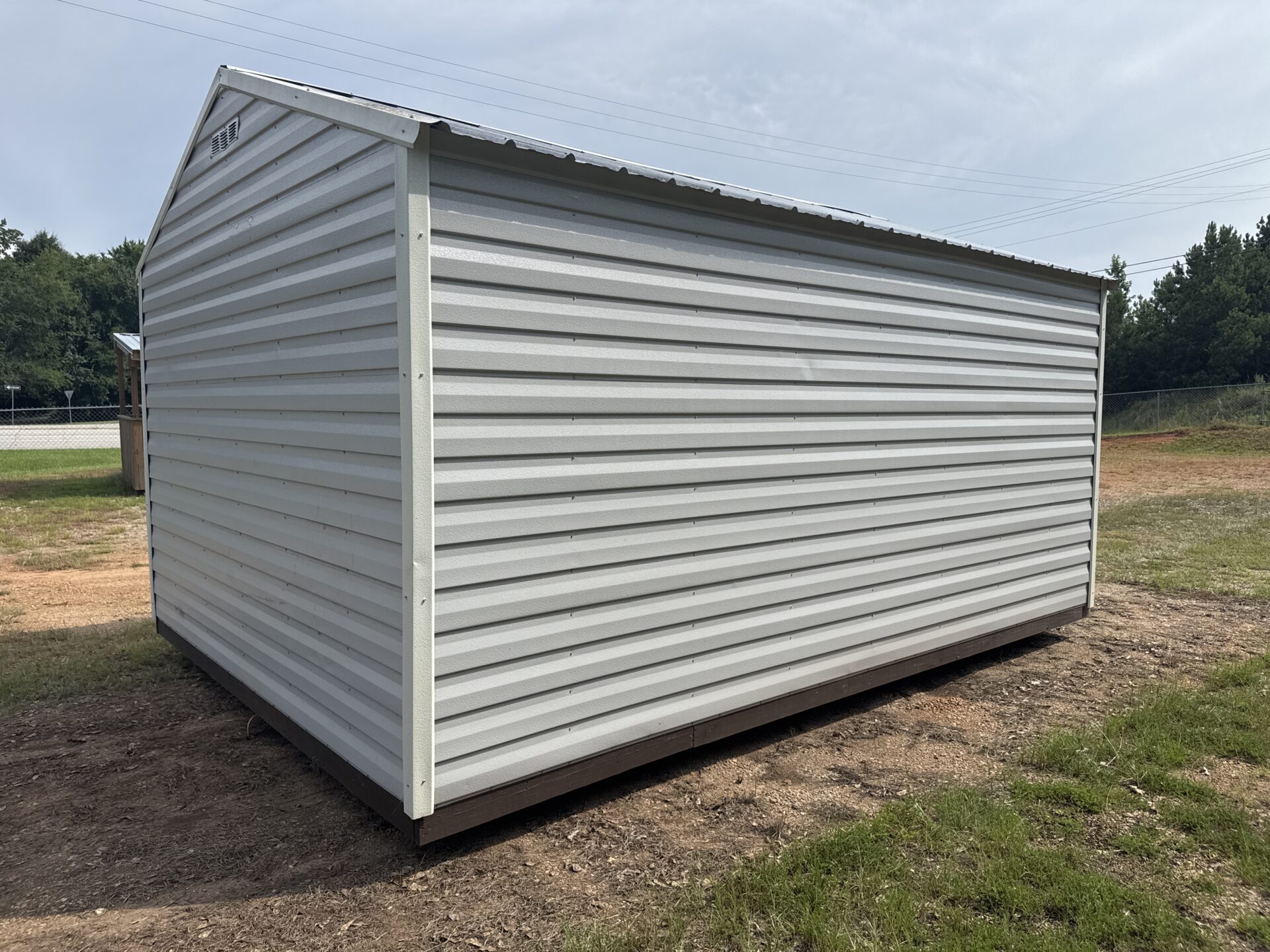 12x16 Ridgeline Used Shed (Stock #071840026) with white metal siding, sloped roof, on bare ground, grass and trees behind.