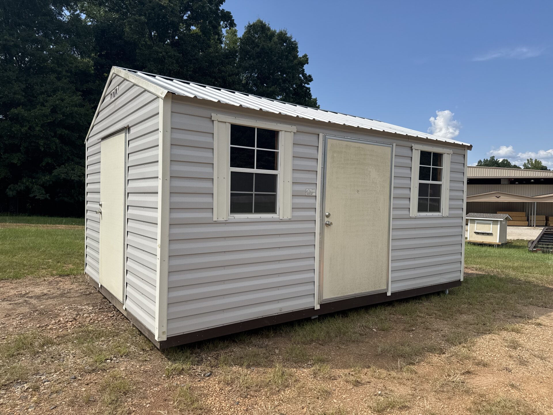 The 12x16 Ridgeline Used Shed with sloped roof, two windows, and a door stands on grass and dirt under a clear sky.
