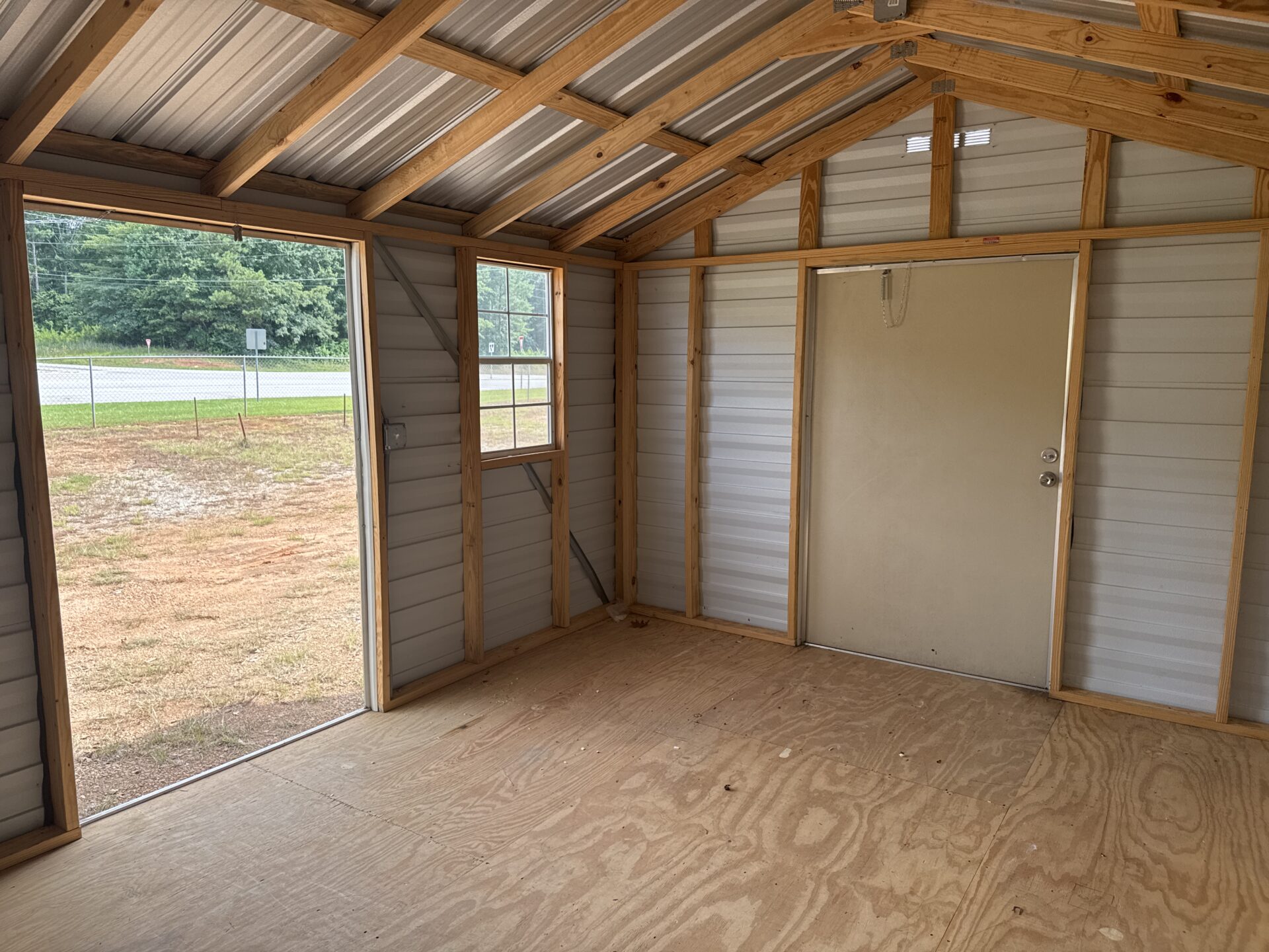 Interior of 12x16 Ridgeline Used Shed with plywood floor, exposed framing, metal roof, open door and window showing trees and road.