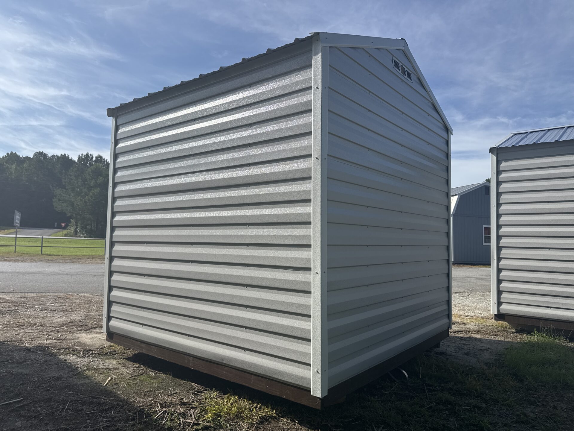 An 8x8 Ridgeline Shed (Stock #071341791), featuring a compact, portable metal design with a sloped roof, sits on a grassy patch beside a road under clear, sunny skies. Another building and trees can be seen in the background.