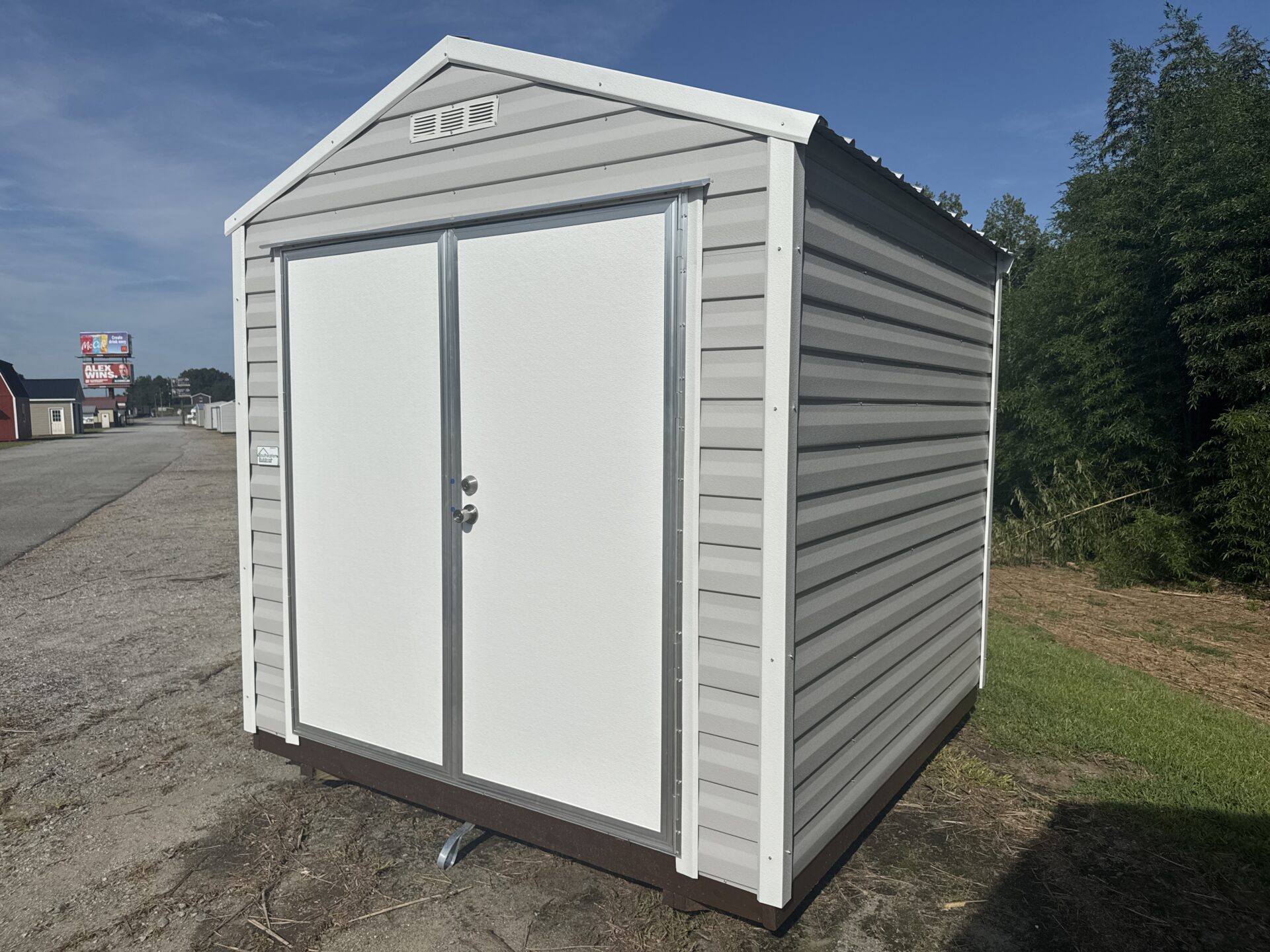 An 8x8 Ridgeline Shed (Stock #071341791) in light gray metal with double white doors is positioned on gravel and grass beneath a clear blue sky. Trees and a roadside sign appear in the background.