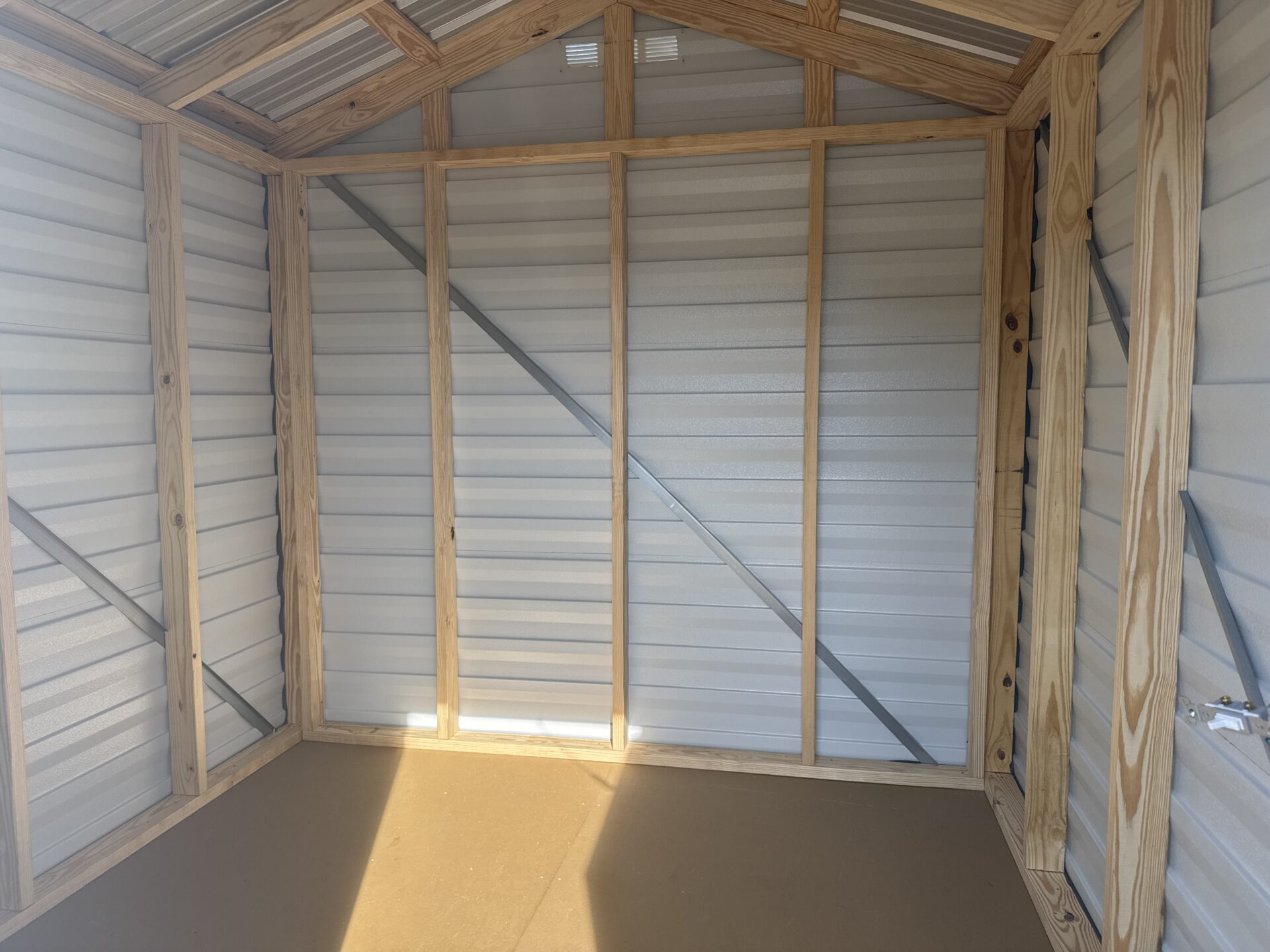 The interior of the 8x8 Ridgeline Shed (Stock #071341791) is shown, with an unfinished wooden frame, metal walls, and sunlight streaming across the floor.