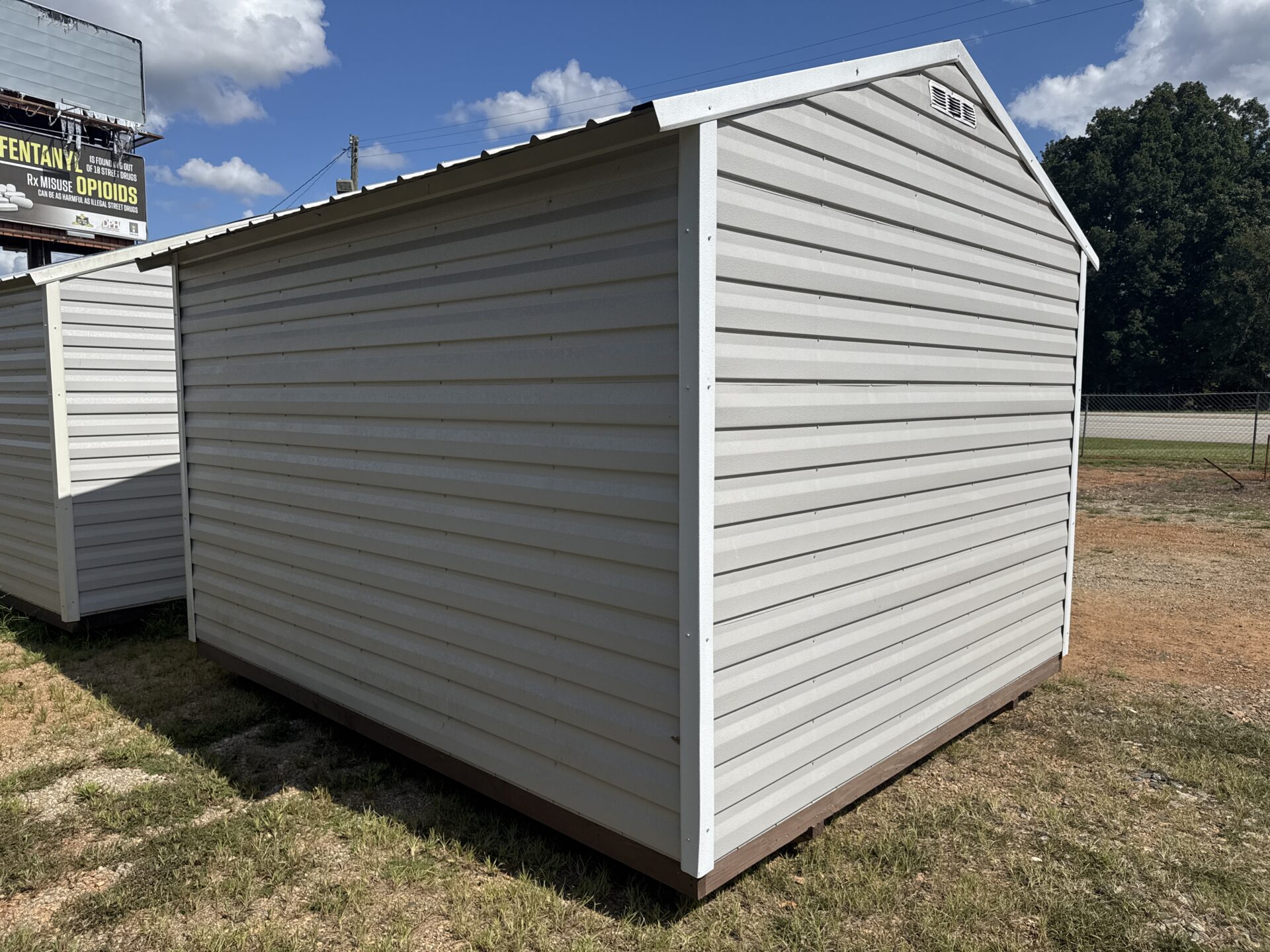 A 10x12 Ridgeline Repo Shed in light gray with white trim sits on a patch of grass and dirt under a sunny sky, with another shed partially visible on the left and a sign in the background. This shed is available now – Stock #032438224.