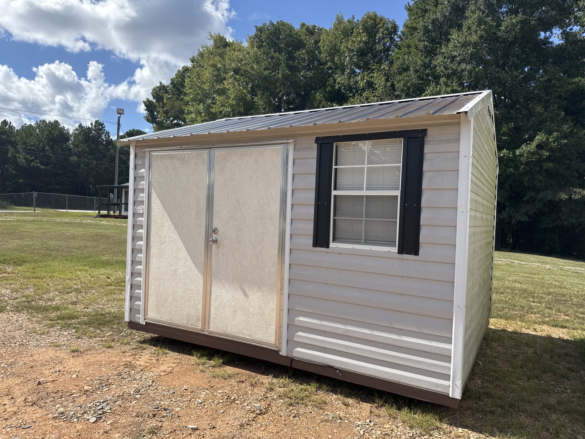 A 10x12 Ridgeline Repo Shed (Stock #032438224), featuring a compact, light gray exterior with double doors and a window accented by black shutters, sits on a dirt patch surrounded by grass and trees under a partly cloudy sky.