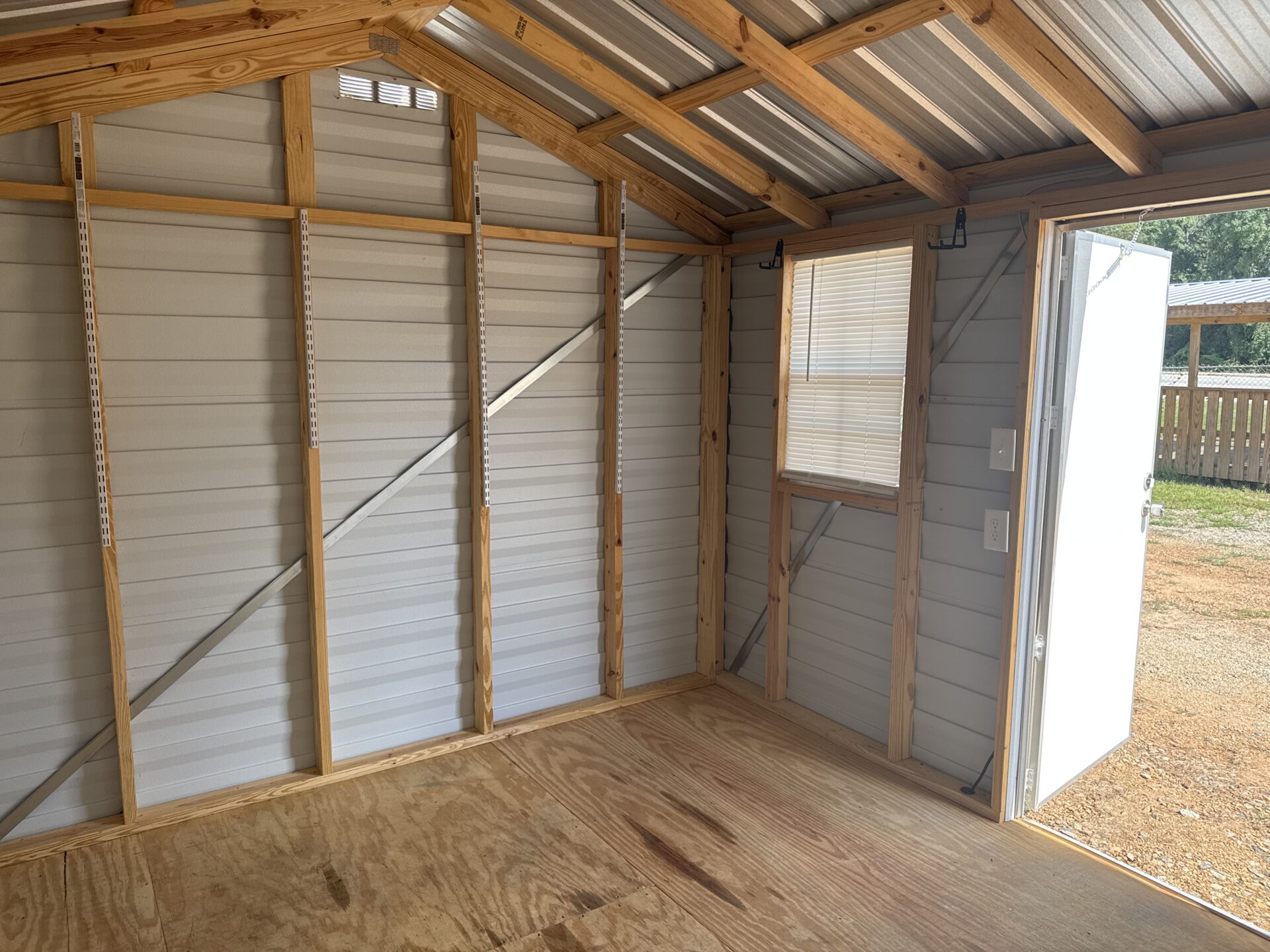 Interior view of the 10x12 Ridgeline Repo Shed, stock number 032438224, showing metal walls, a window, wall-mounted hooks, a plywood floor, and an open door with sunlight streaming in. Now available.