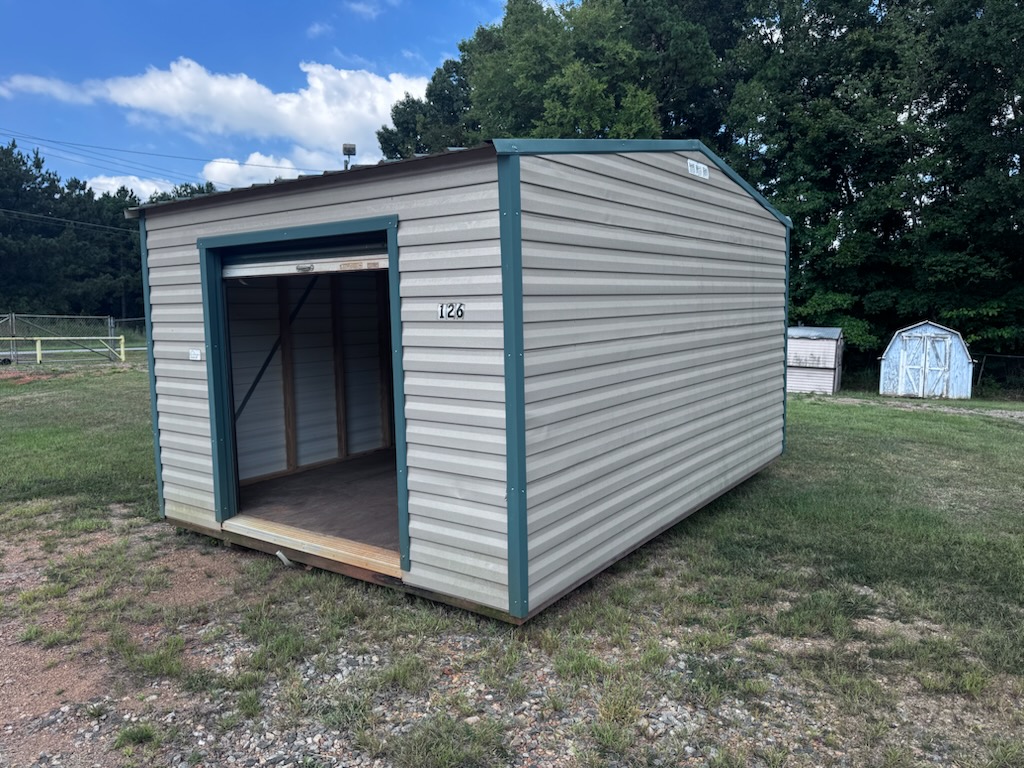 A 12x16 Storage Garage Shed – Scratch & Dent, Stock #0622EP30538, sits on grass with its door partially open. The light gray shed is surrounded by trees and other small sheds under a partly cloudy sky.