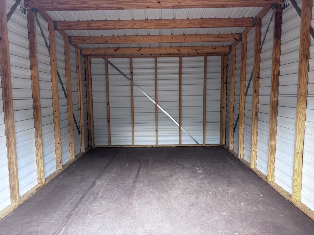 Interior of a 12x16 Storage Garage Shed (Stock #0622EP30538) featuring an empty space with wooden framing, a brown floor, and white metal walls and roof. The well-lit area is clean, shows exposed beams, and has a simple rectangular layout. This Scratch & Dent shed is available now.